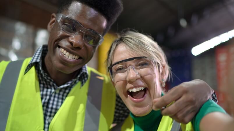 Young man and woman with yellow safety vests and glasses, large smiles, arm in arm